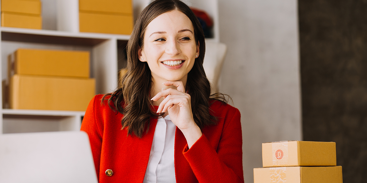 young woman in red blazer sits at computer with packing boxes